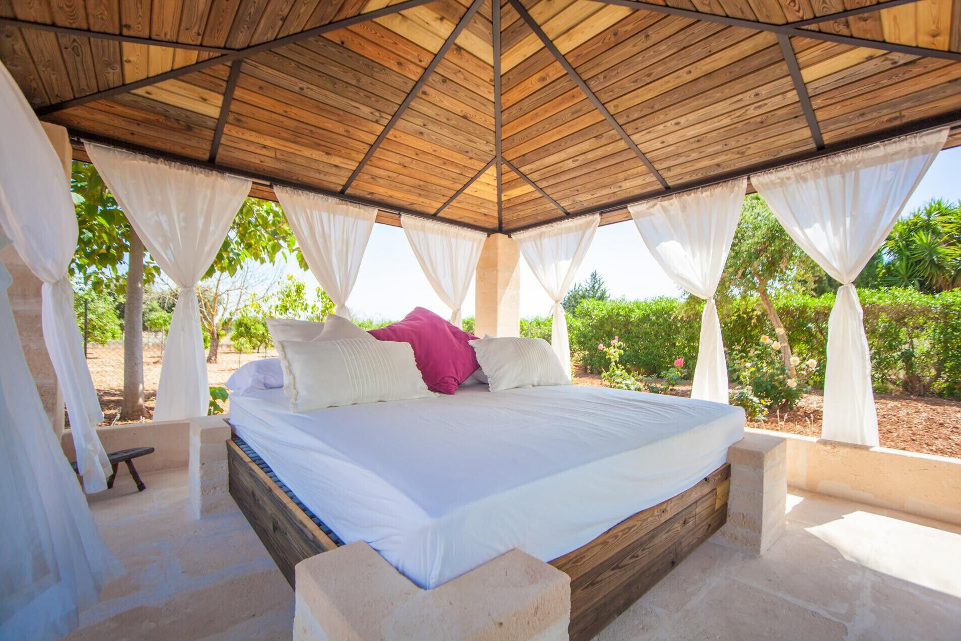 Un lit avec une literie blanche et un oreiller rose, situé dans un espace extérieur abrité, doté de poutres en bois et de rideaux blancs.