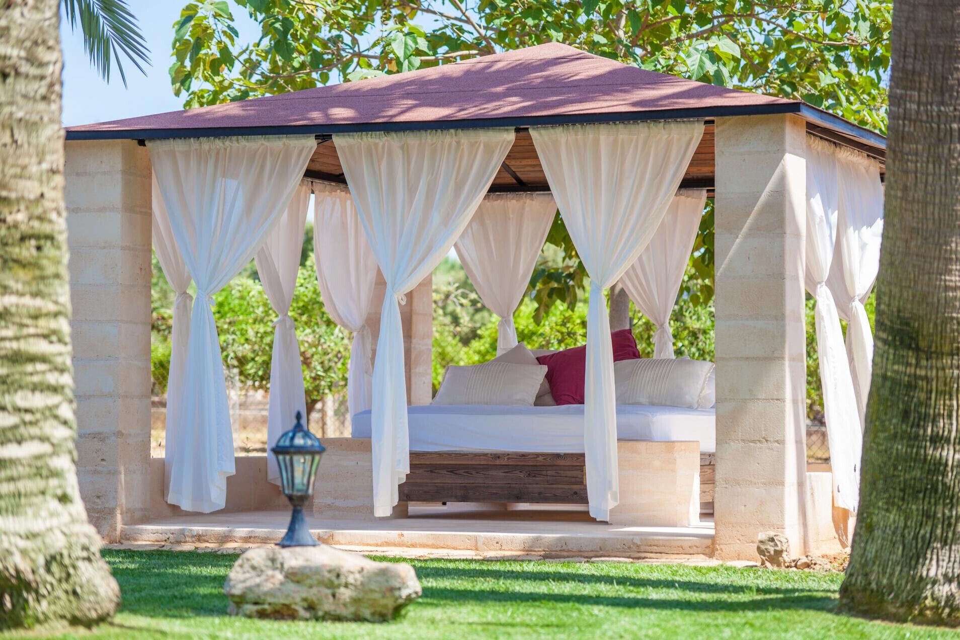 Une chambre à coucher extérieure de style gazebo, avec un lit, des rideaux blancs et une lanterne.
