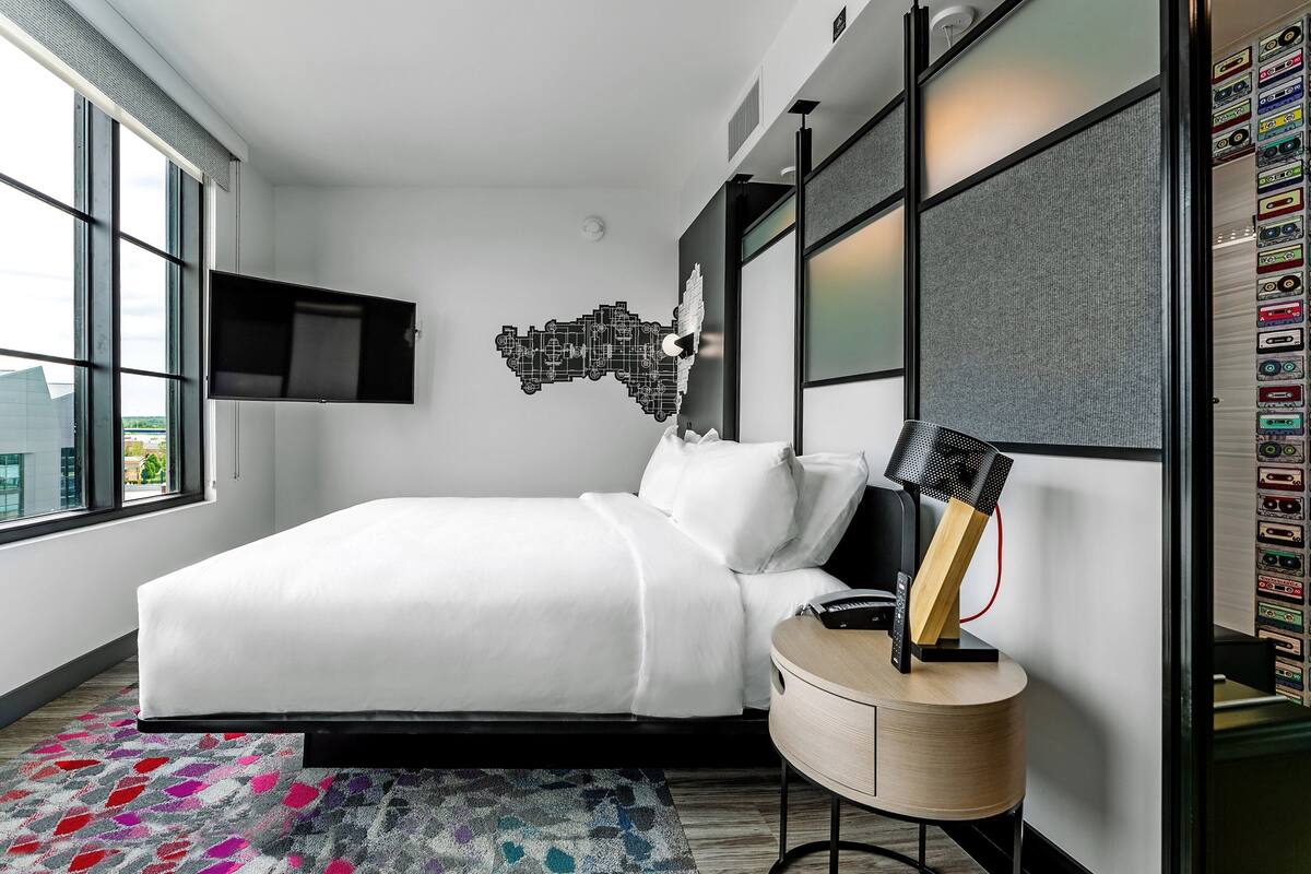 A modern hotel room with a large bed, a mounted TV, a unique wall art piece, and a small side table with a lamp.