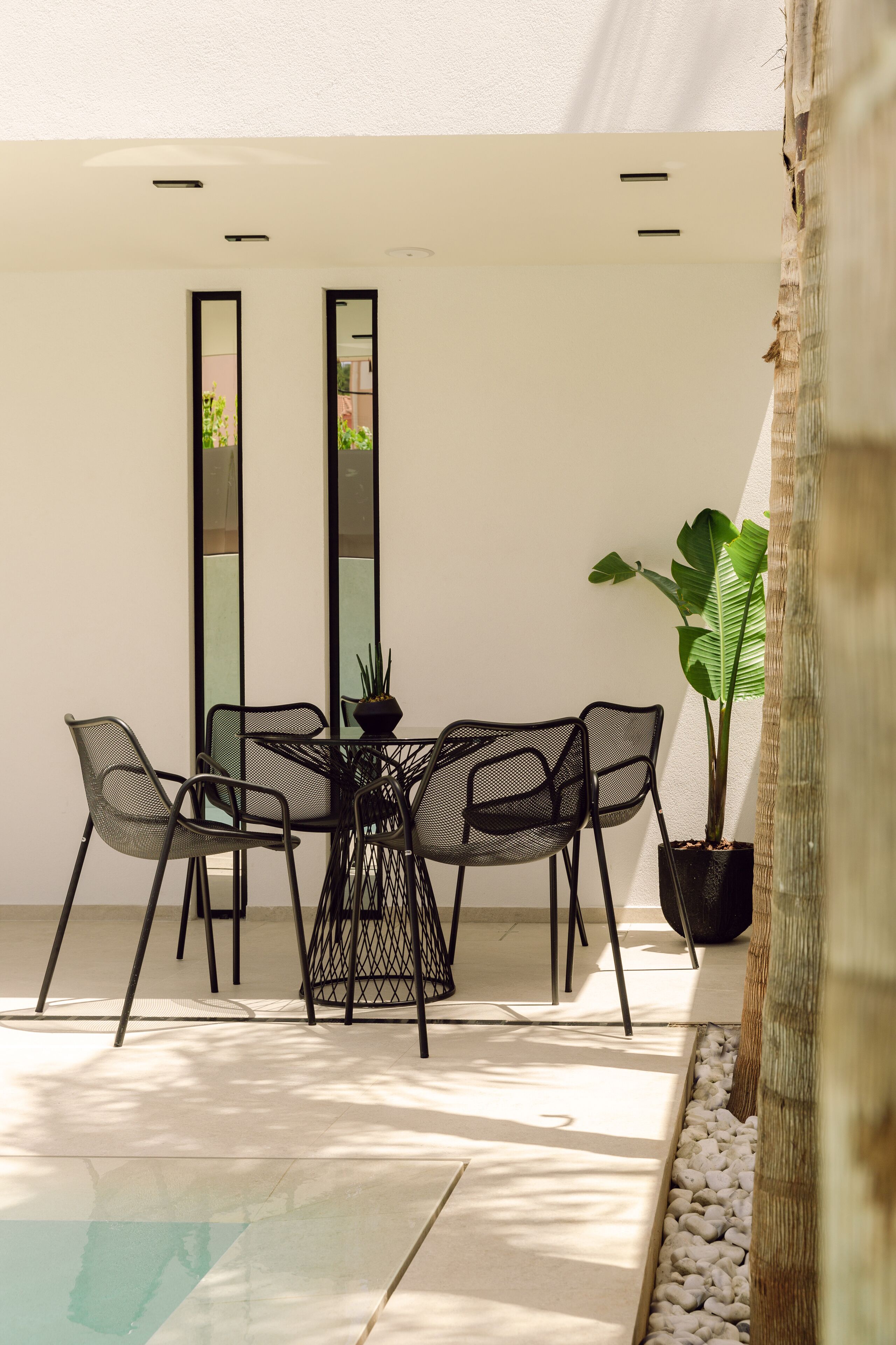 A modern outdoor dining area with a round table and chairs, a potted plant, and a view of a building through a window.