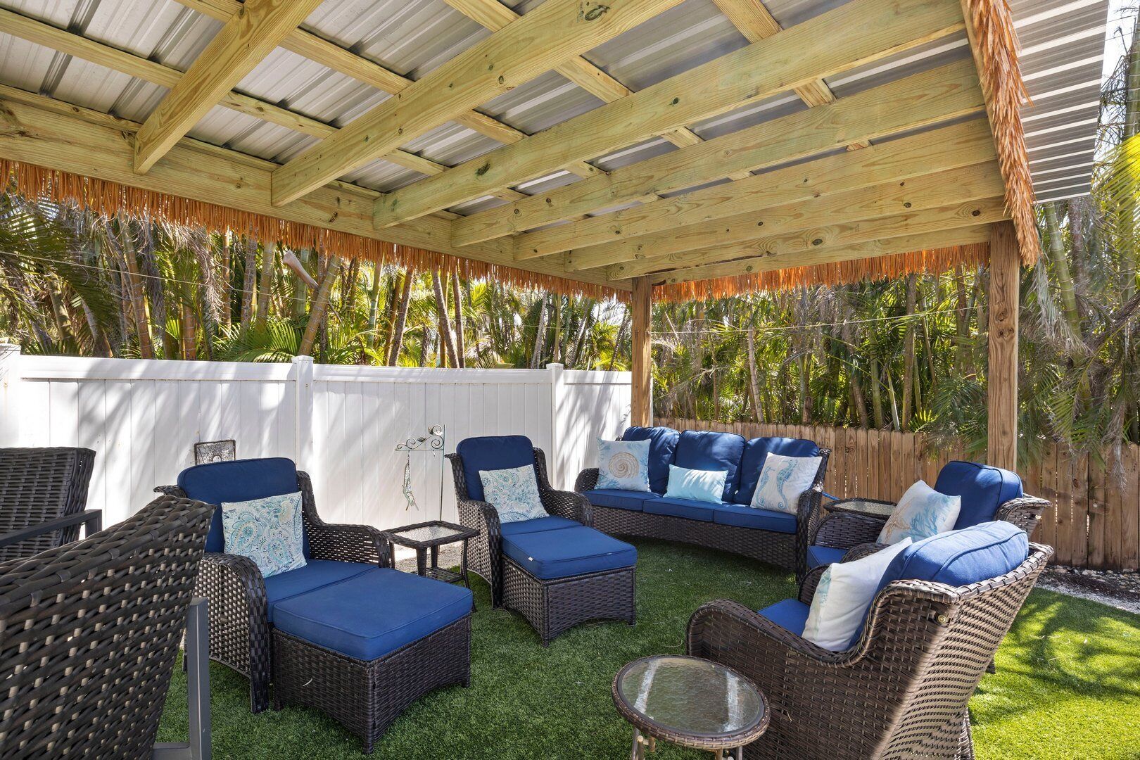 Un espace extérieur aménagé, abrité et doté de mobilier en osier et de coussins bleus, entouré d’une clôture blanche et de plantes tropicales.