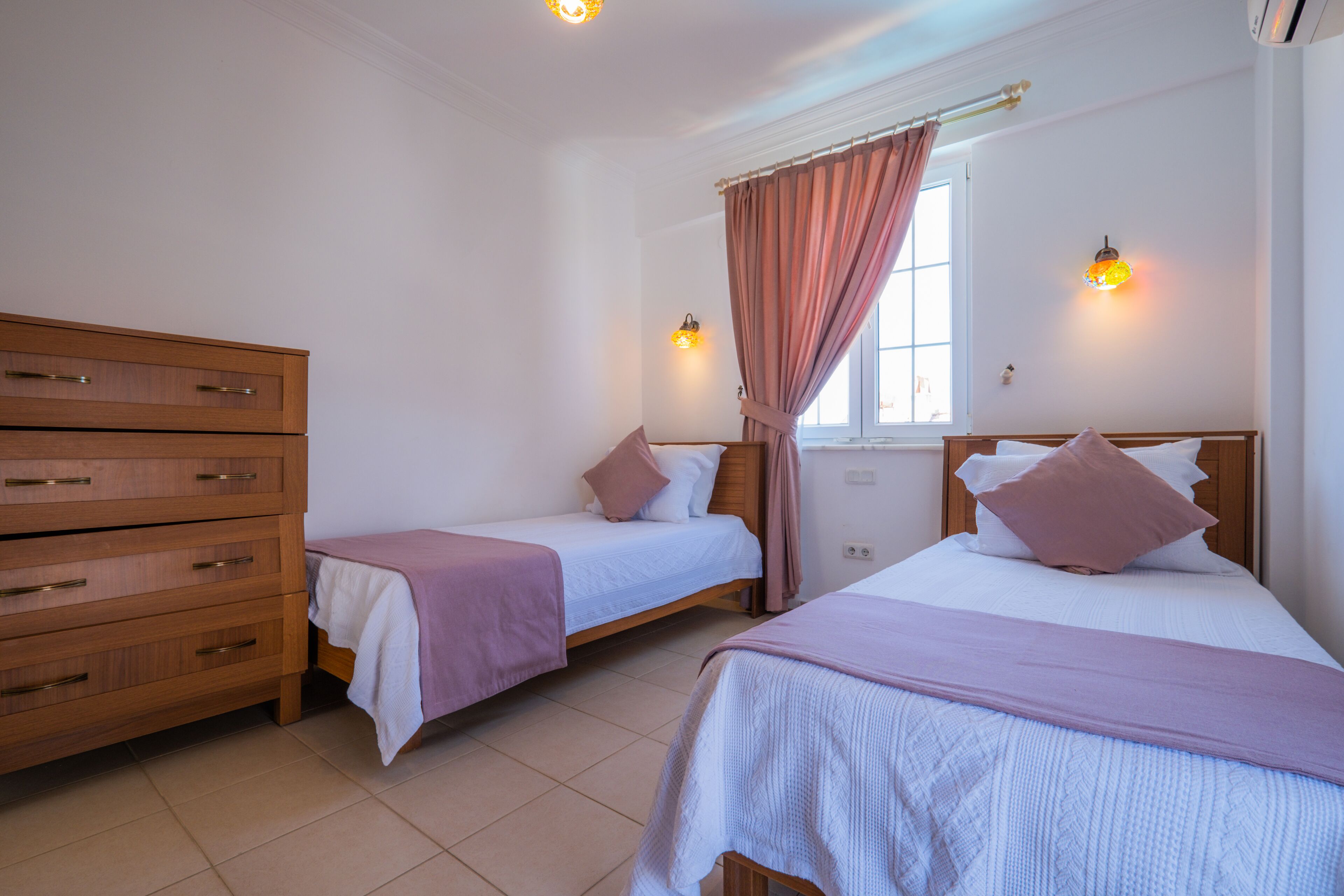 A bedroom with two single beds, a wooden dresser, and a window with curtains.