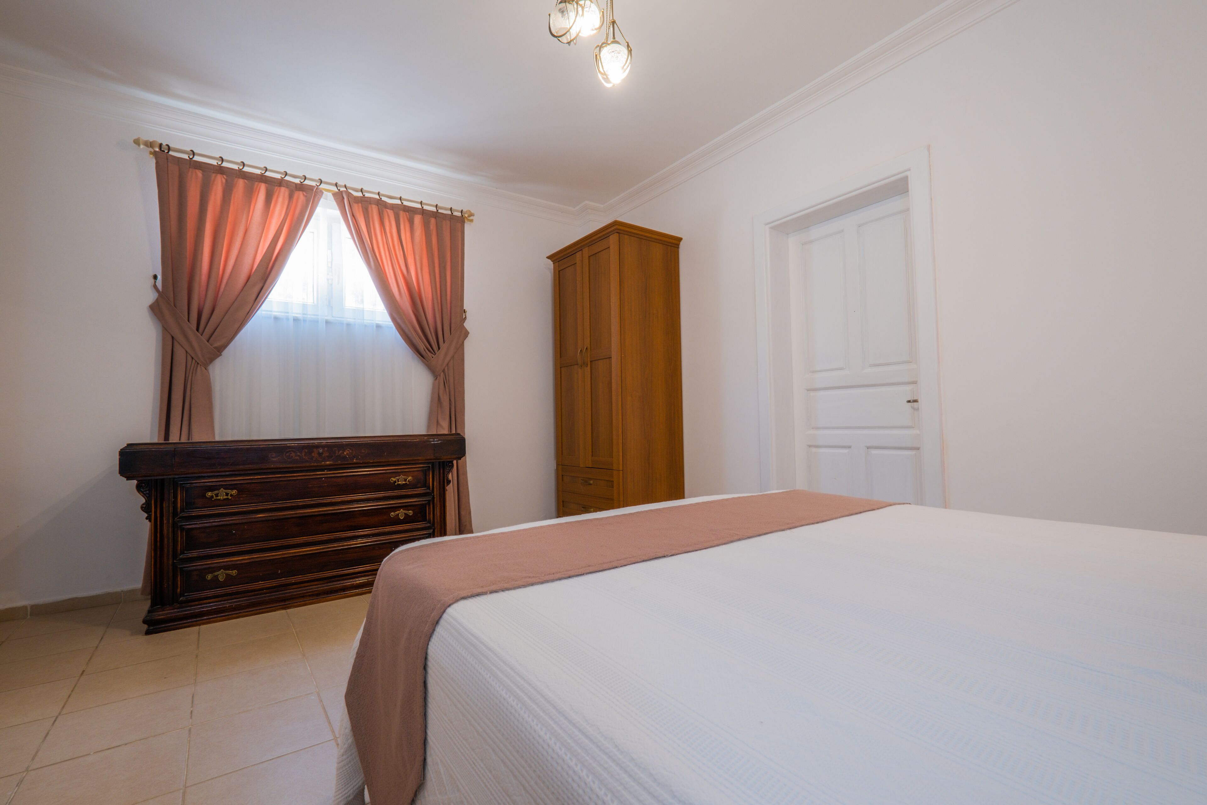 Un dormitorio con cama, armario de madera, cómoda y ventana con cortinas.