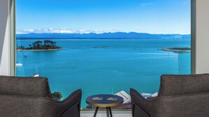 A room with a large window overlooking a turquoise sea, two chairs, and a small round table.