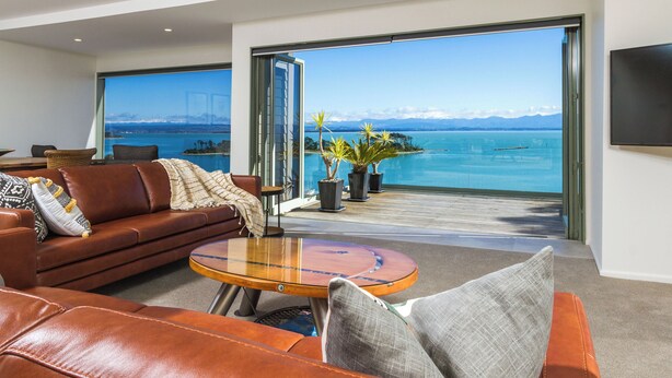 A modern living room with a leather sofa, a round coffee table, and a large sliding glass door leading to a balcony with a view of the sea and mountains.