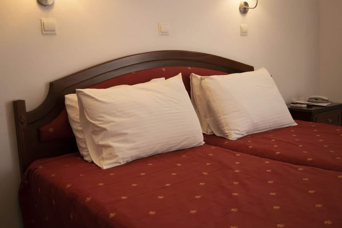Een tweepersoonsbed met een houten hoofdbord, twee witte kussens en een rode sprei met een subtiel patroon.