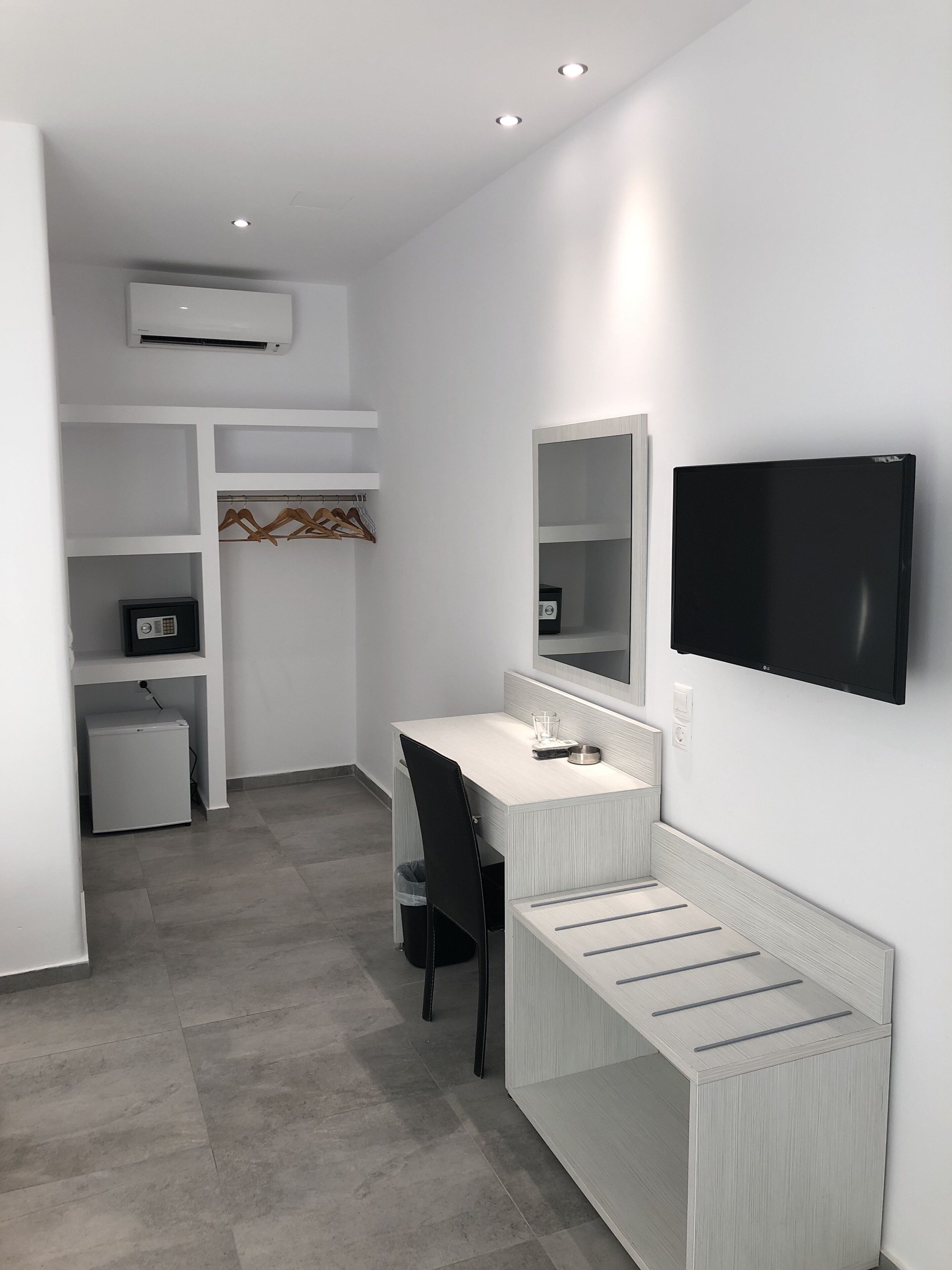 A modern, minimalistic room with a white desk, a black chair, a TV mounted on the wall, and a small refrigerator.