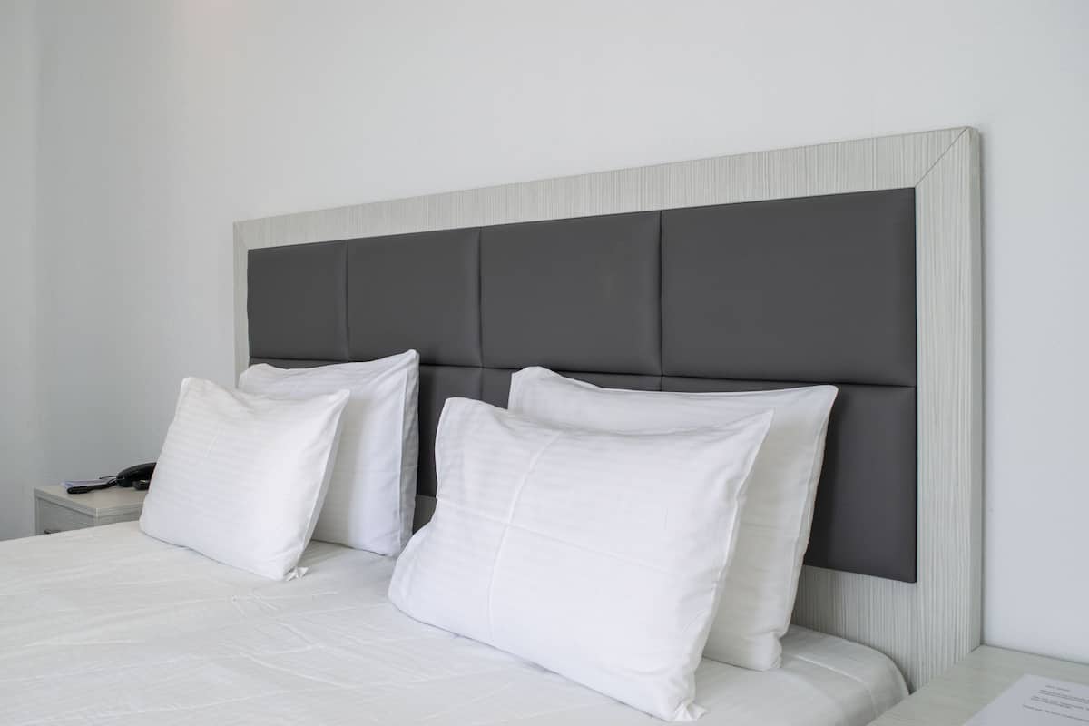 Een net opgemaakt bed met witte beddengoed en kussens, een grijs gestoffeerd hoofdbord, en een witte nachtkastje met een lamp.