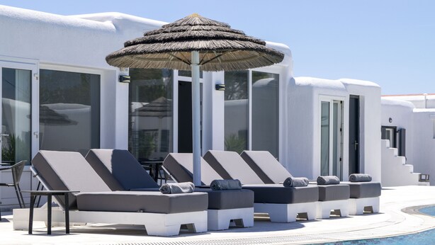 Un espace aménag é au bord de la piscine, avec des chaises longues et un parasol en chaume, entouré de bâtiments blancs dotés de grandes fenêtres.