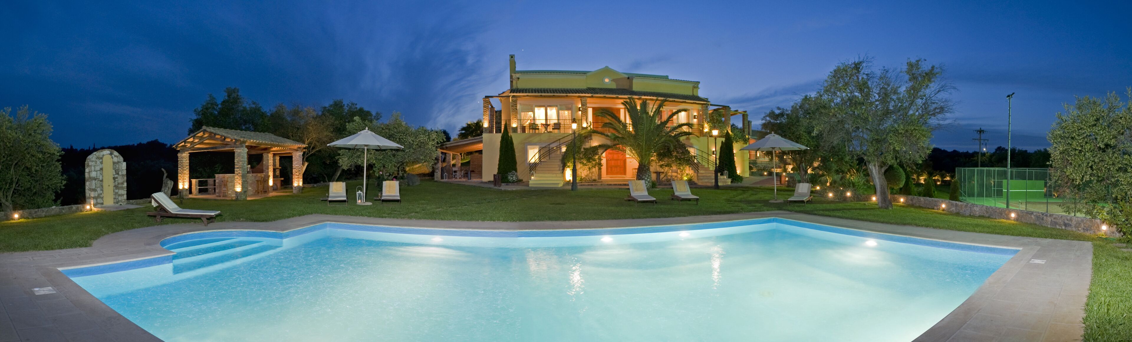 A large, well-lit swimming pool with lounge chairs and umbrellas, surrounded by a landscaped garden and a large house with multiple levels and outdoor area lighting.