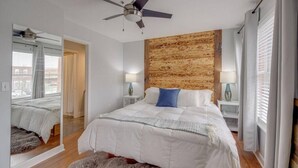 Un dormitorio con una cama grande, un ventilador de techo, cabecera de madera, dos mesitas de noche con lámparas, un espejo y vistas al exterior a través de una ventana.