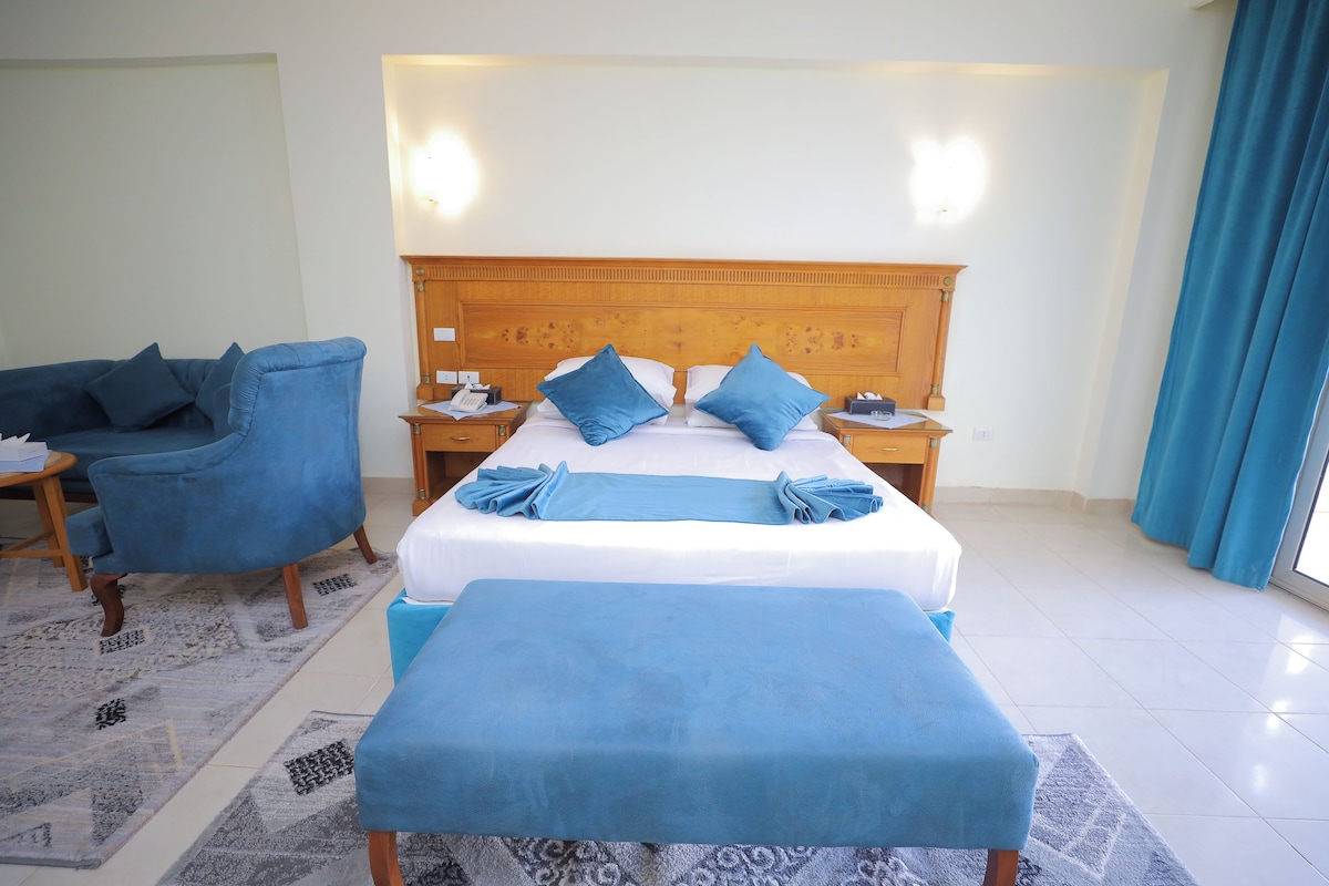Una camera d'hotel con un letto grande, due comodini, una poltrona blu e una panca.