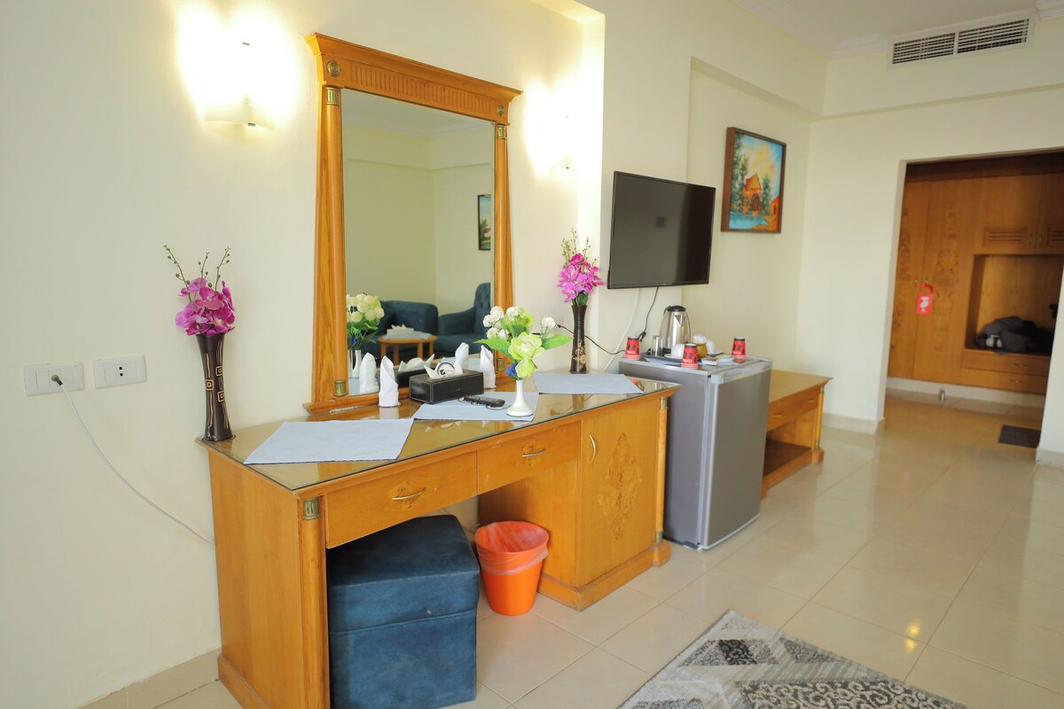 A hotel room with a wooden desk, a mirror, a television, and a small kitchenette.