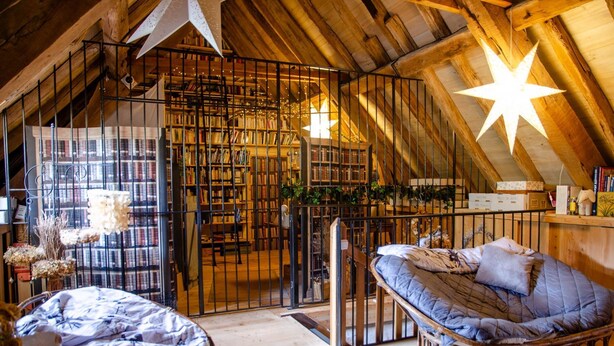 Een gezellige zolderkamer met een hoge plafond, houten balken, een ster-vormige lamp, een bed, een stoel en een boekenplank vol boeken.