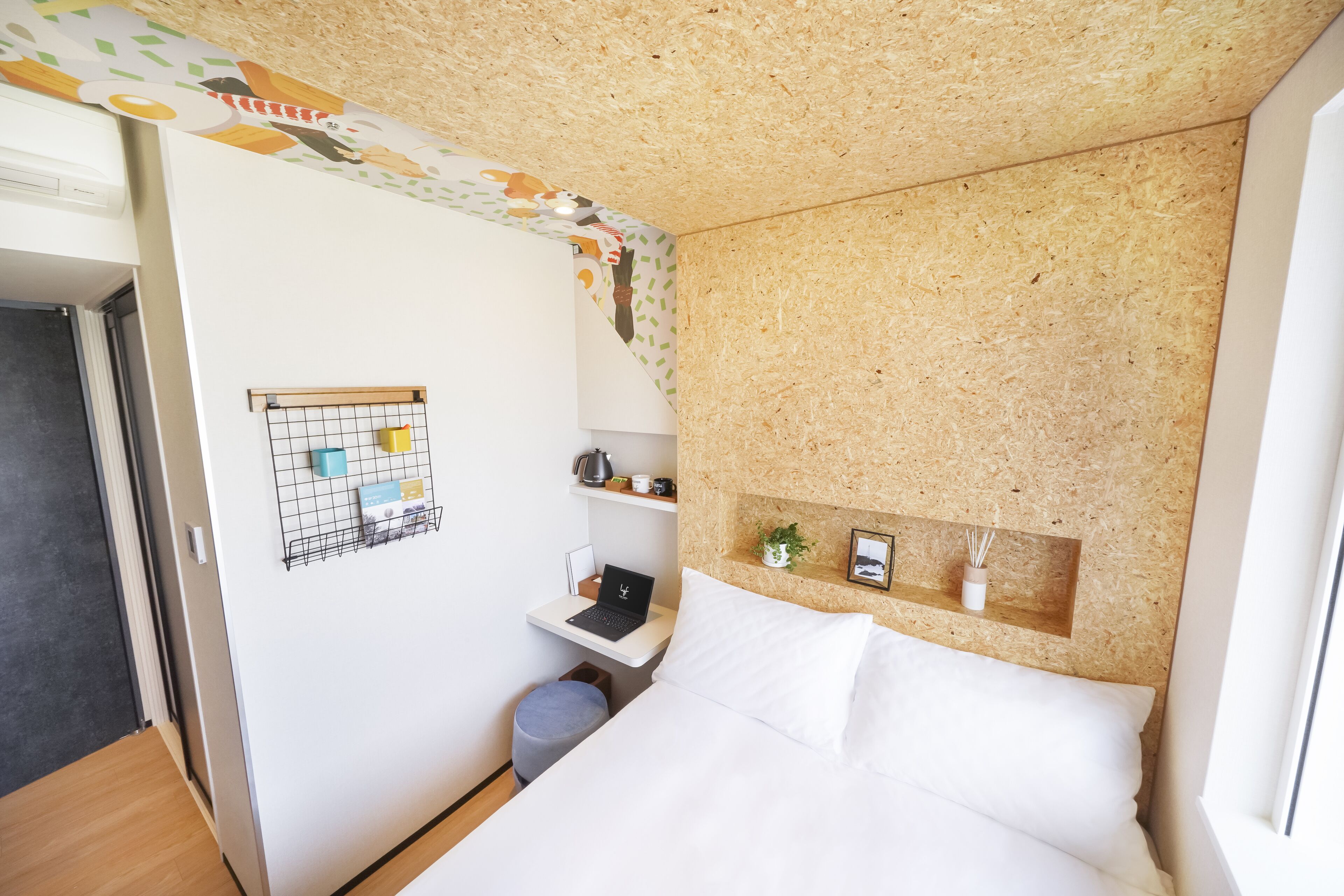 Ein kleines Zimmer mit einem Bett, einem Schreibtisch mit einem Laptop, einem kleinen Hocker und einem Regal mit persönlichen Gegenständen.