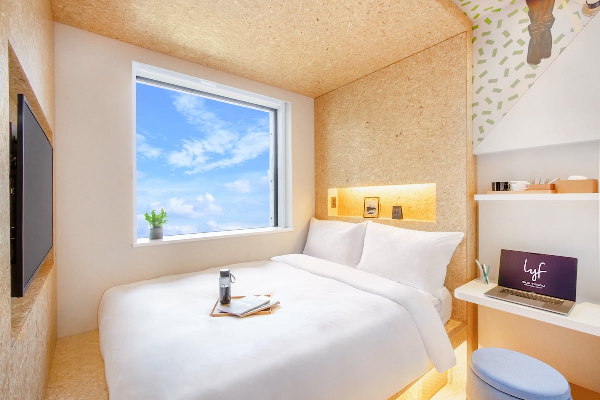 Moderni hotellihuone, jossa on suuri sänky, työpöytä kannettavalla tietokoneella, ikkuna, josta avautuu näkymä taivaalle, ja pieni kasvi ikkunalaudalla.