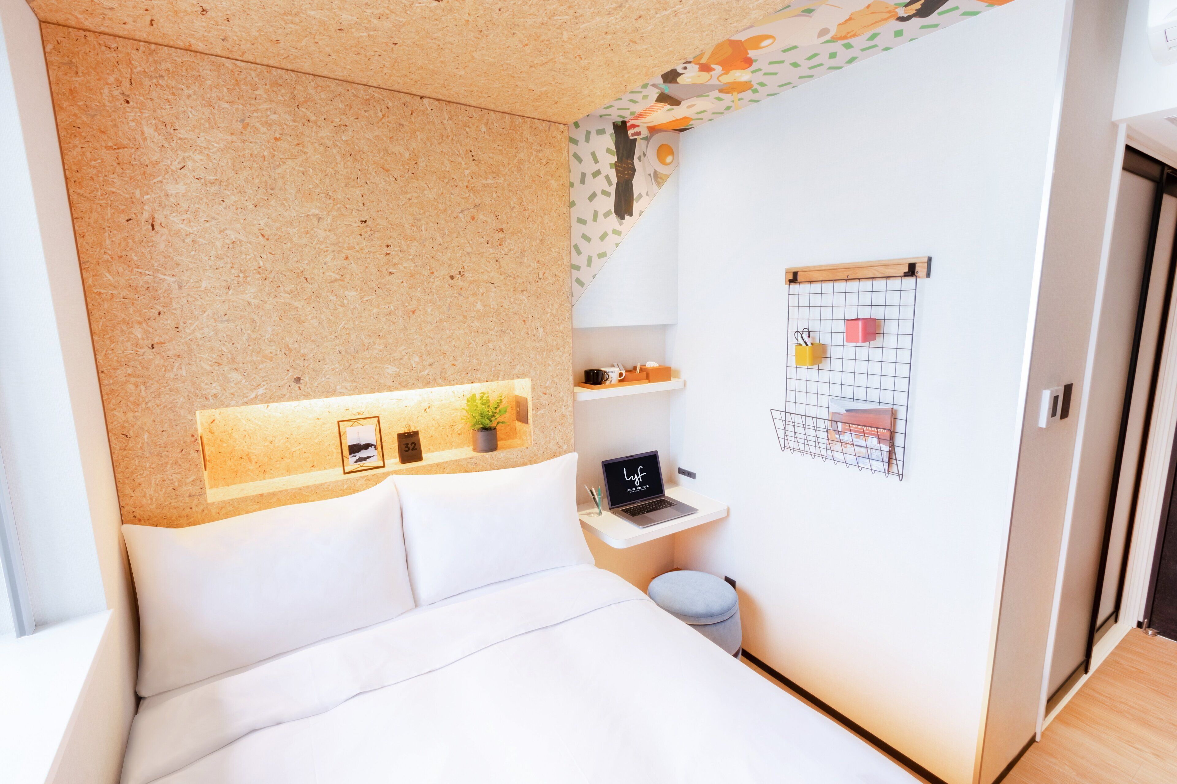 Ein Schlafzimmer mit einem Bett, einem kleinen Schreibtisch mit einem Laptop, einer Zimmerpflanze und einem Wandkalender.