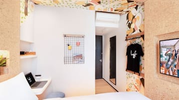 A modern hotel room with a bed, a desk with a laptop, a coffee cup, a small plant, and a wall-mounted shelf.