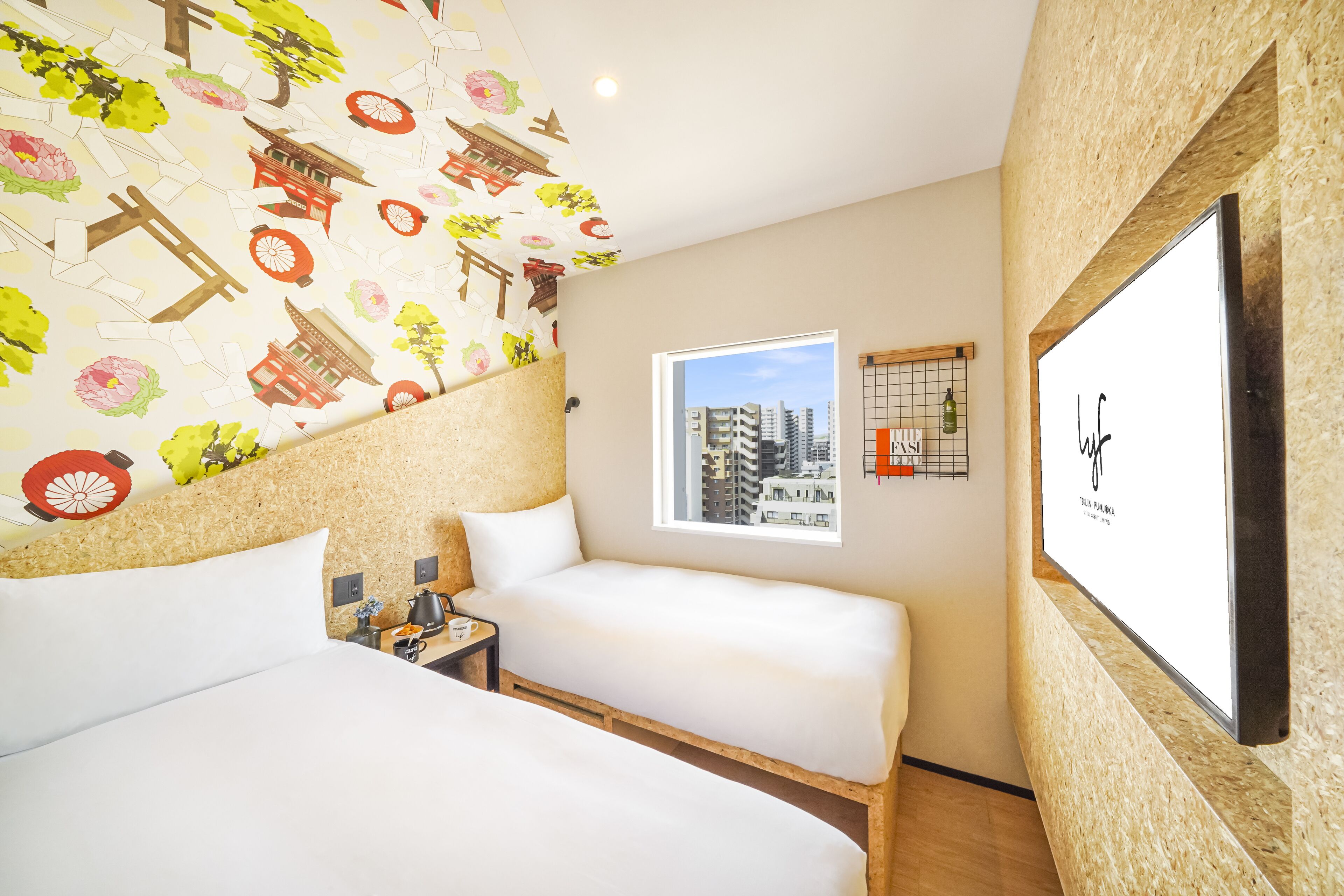 Una habitación de hotel con dos camas, un ventanal con vista a la ciudad, un televisor montado en la pared y una pared decorativa con motivos culturales.