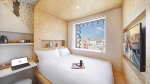 Une chambre d’hôtel compacte comprenant un lit, un ordinateur portable posé sur une étagère, une fenêtre donnant sur la ville et un téléviseur fixé au mur.