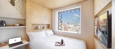 A compact hotel room with a bed, a laptop on a shelf, a window with a city view, and a mounted TV.