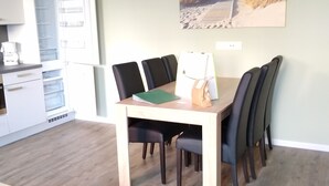 Un comedor con mesa y sillas de madera, una cocina con armarios blancos y un refrigerador, y un cuadro de una playa en la pared.
