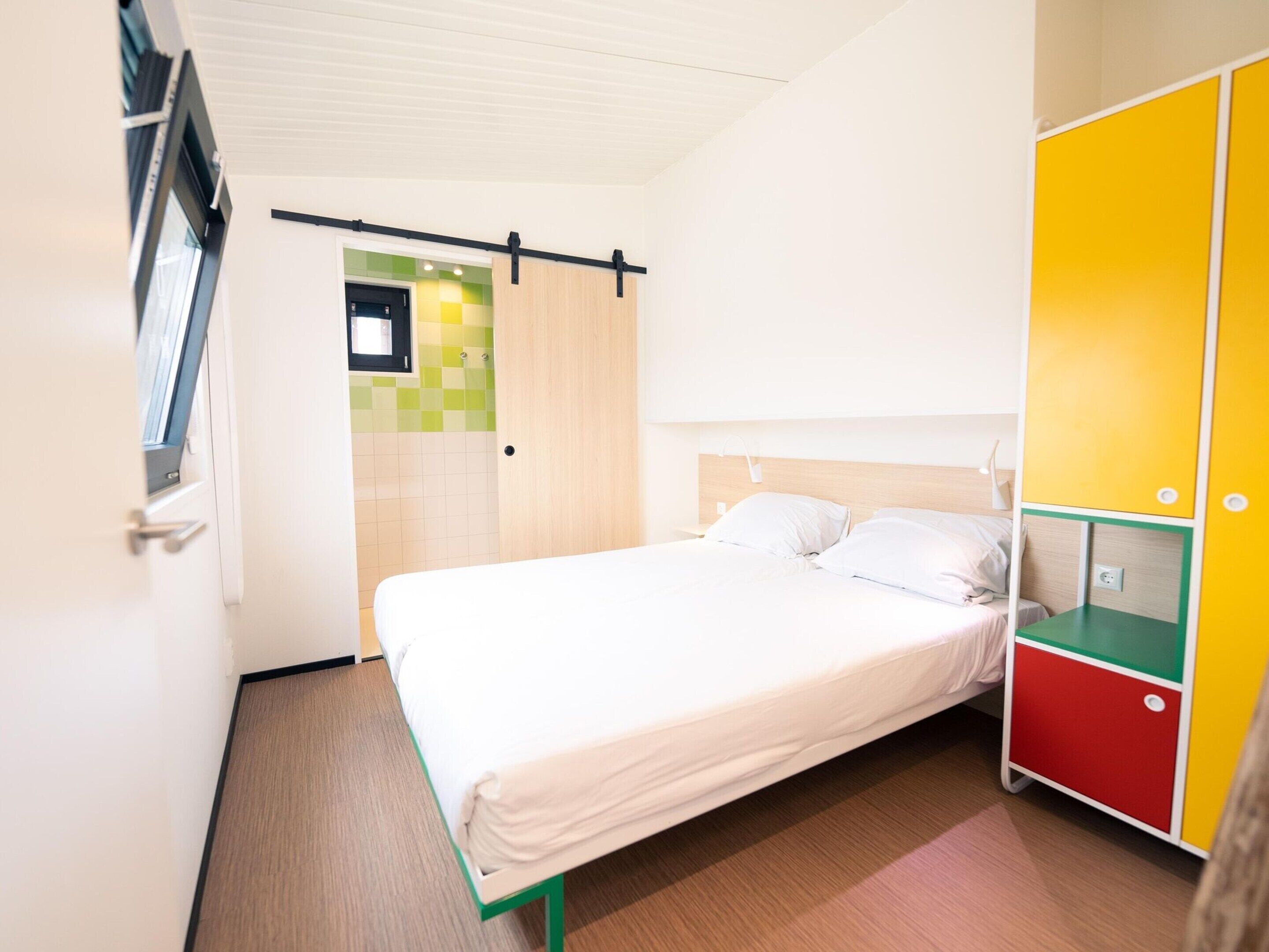 Une chambre avec un lit simple, une tête de lit en bois, une table de chevet et une armoire dont la façade est décorée de motifs colorés.