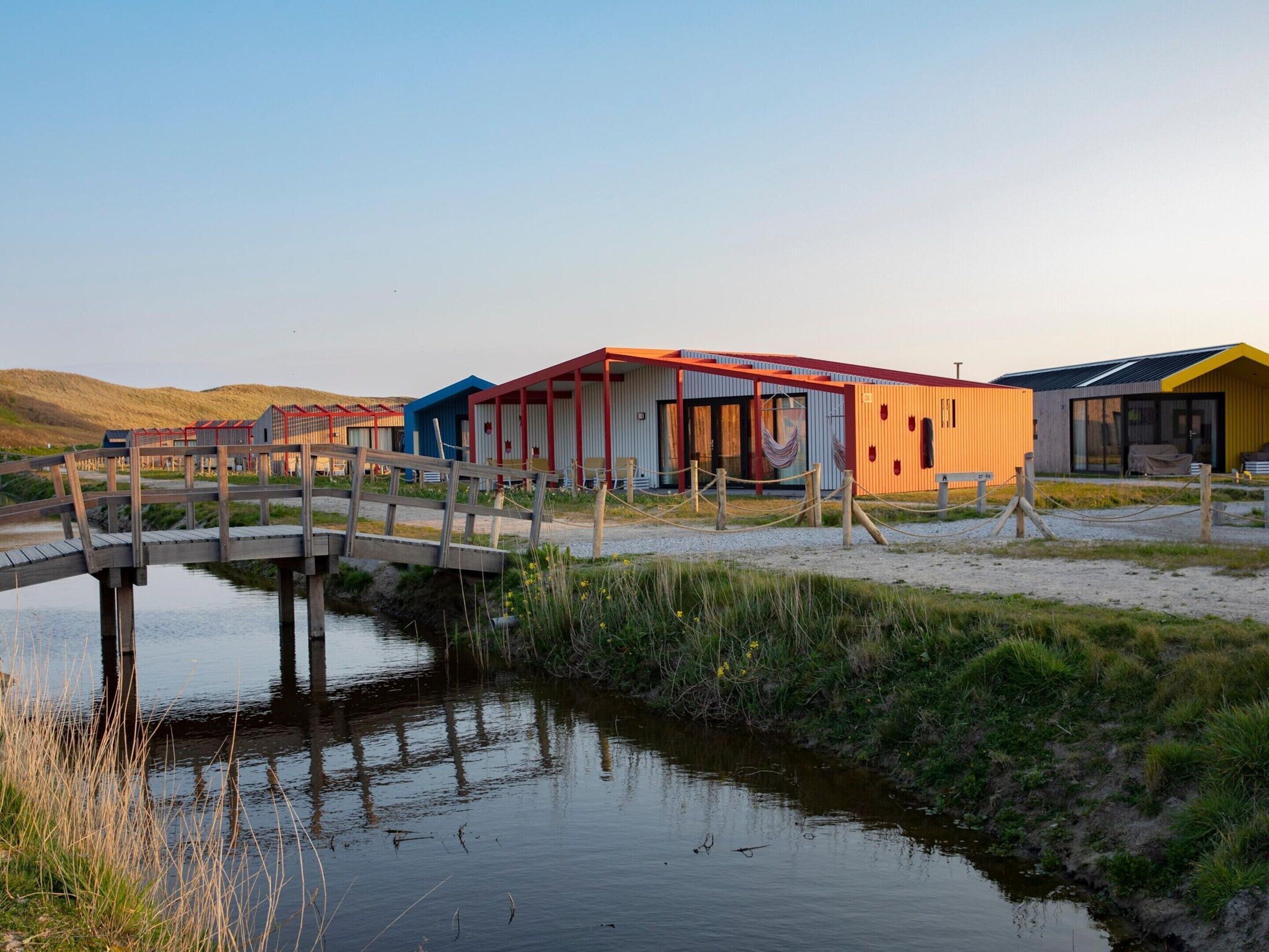 A series of colorful, modern buildings with a wooden bridge over a calm water body, surrounded by grassy areas.