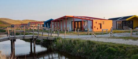 A series of colorful, modern buildings with a wooden bridge over a calm water body, surrounded by grassy areas.