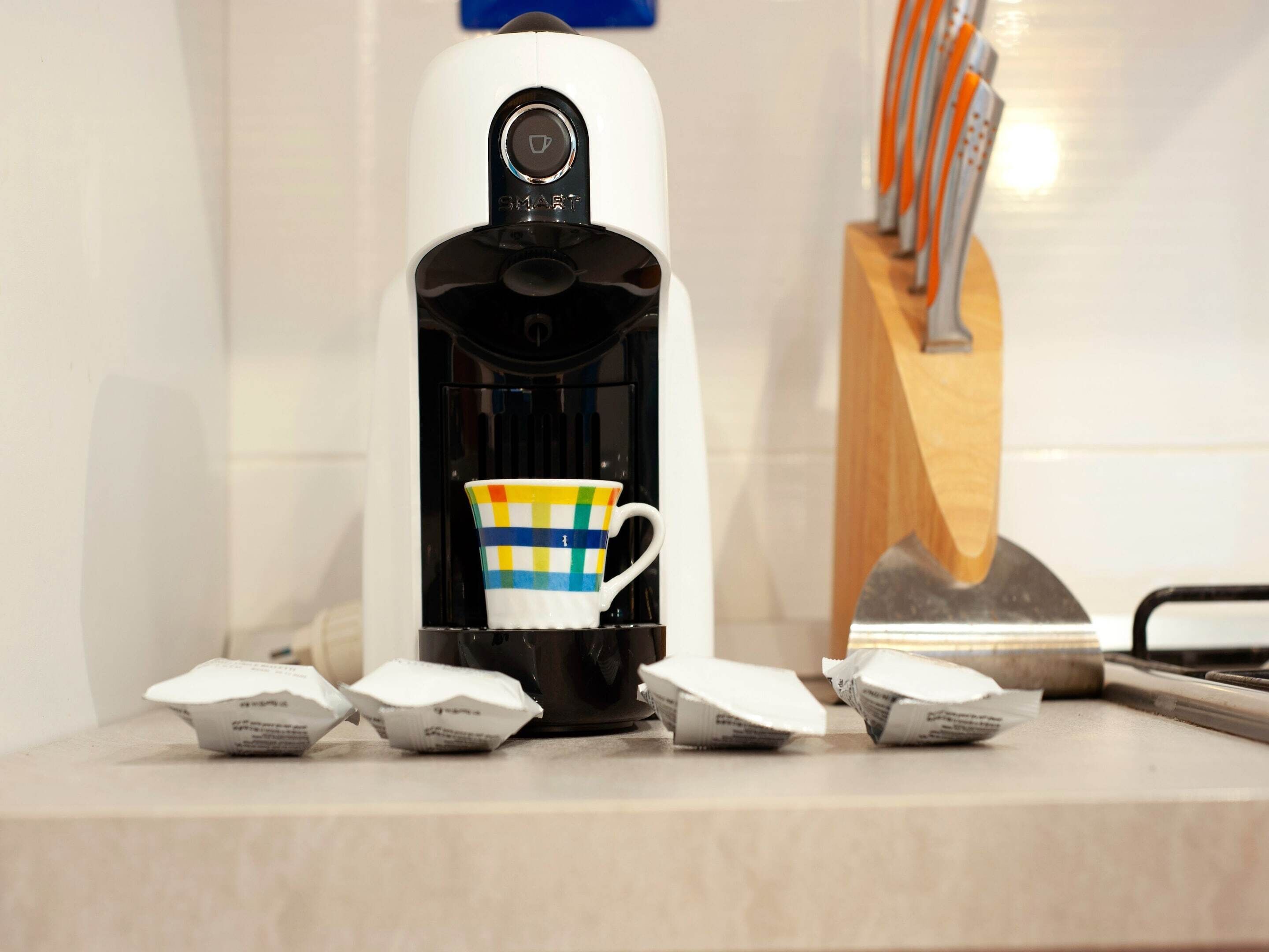 Una cafetera, una taza de colores, un bloque de cuchillos y cápsulas de café sobre una encimera de cocina.
