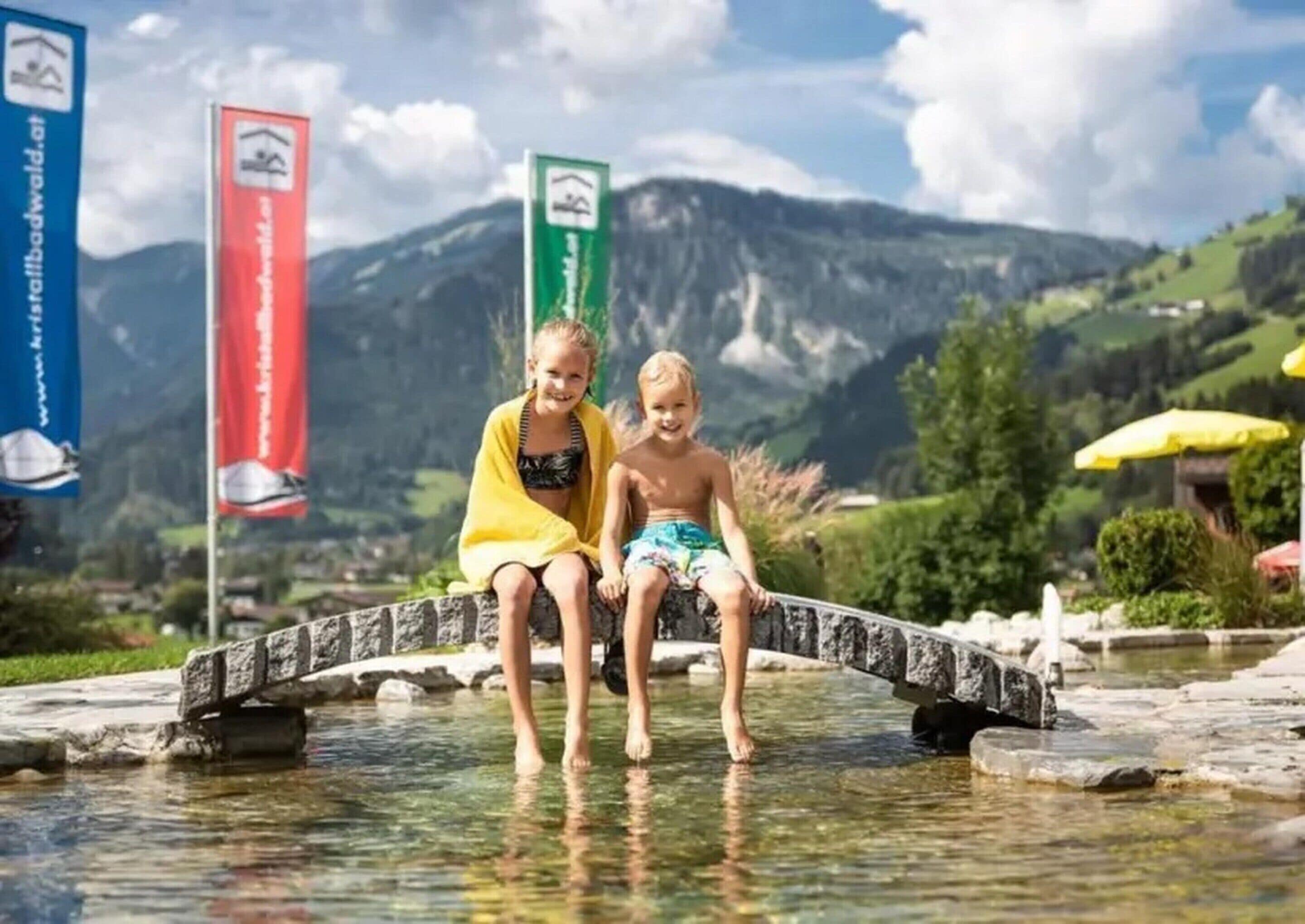 Deux enfants sont assis sur un pont de pierre qui enjambe un cours d’eau limpide, avec des montagnes en arrière-plan.