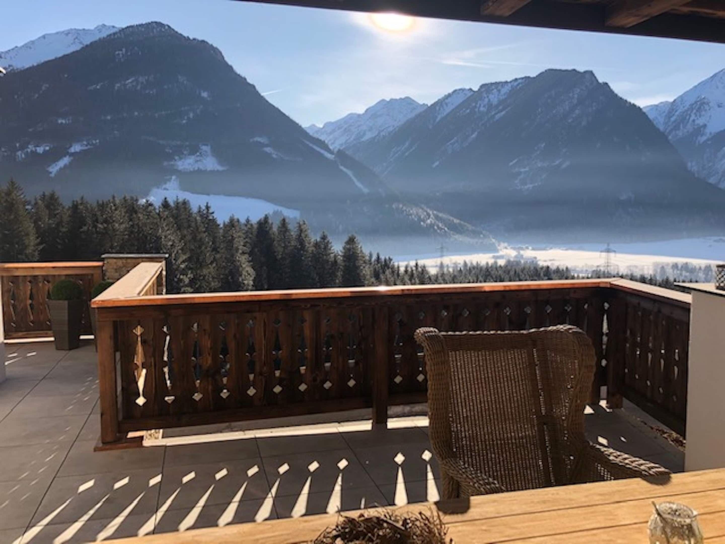 Um deck de madeira com uma cadeira de vime e vista para montanhas nevadas e um lago.
