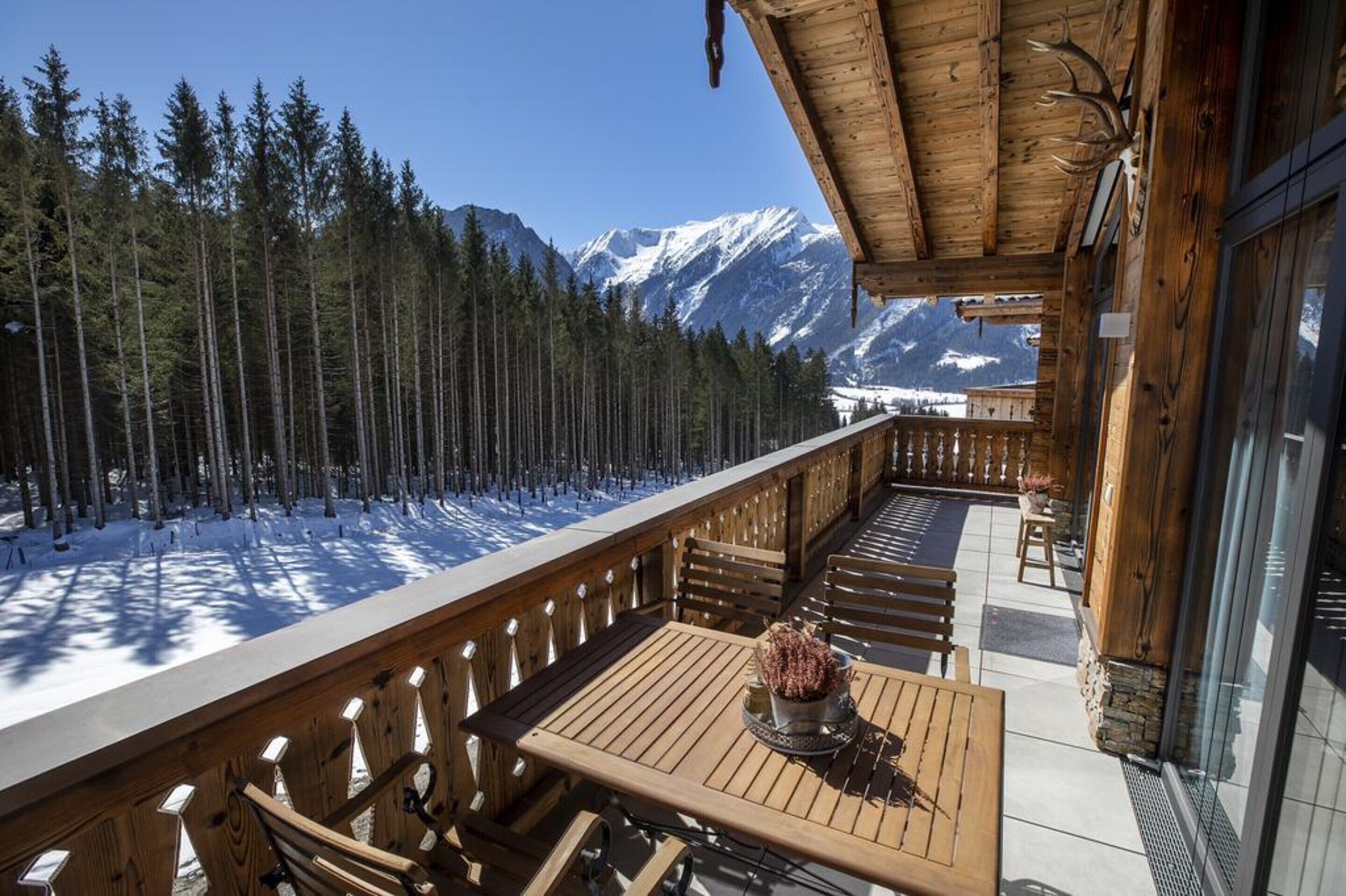Une terrasse en bois avec une table et des chaises, offrant une vue sur une forêt enneigée et des montagnes.