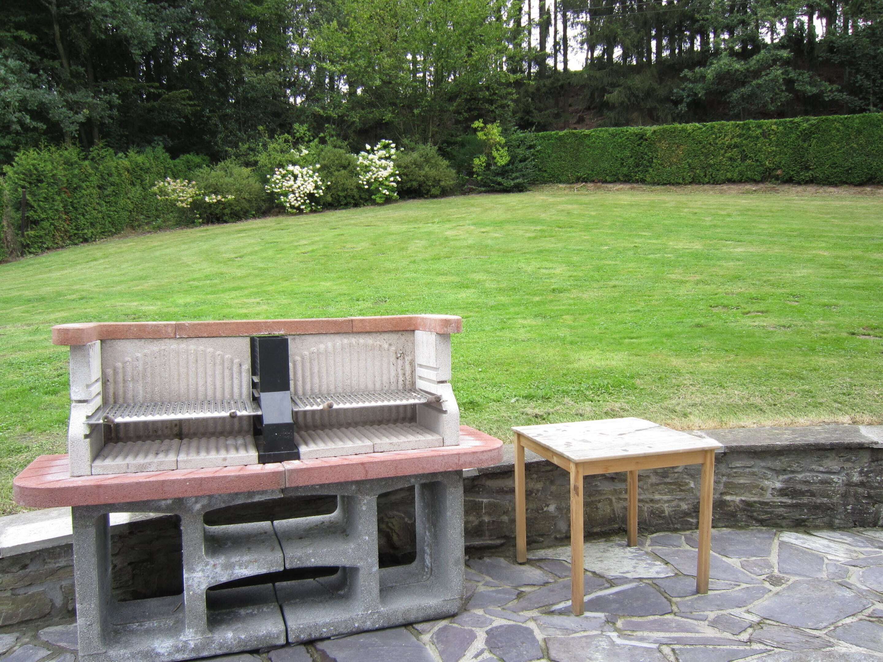 Una parrilla de hormigón con una mesa de madera a un lado, ubicada en un patio empedrado con césped y una siega al fondo.