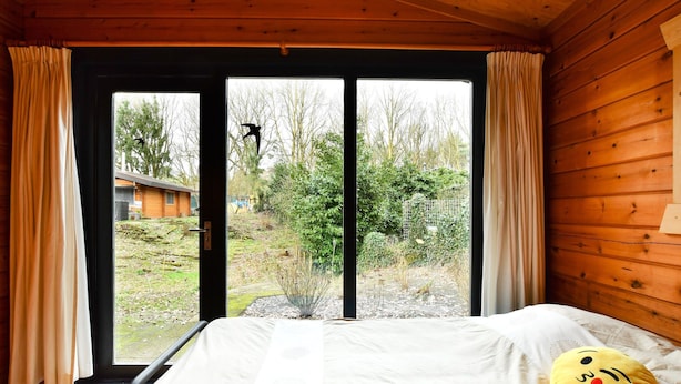 A wooden cabin room with a bed, a window with curtains, and a view of a garden. - Wooden Holiday Home in Meijel With Private Garden (Meijel)