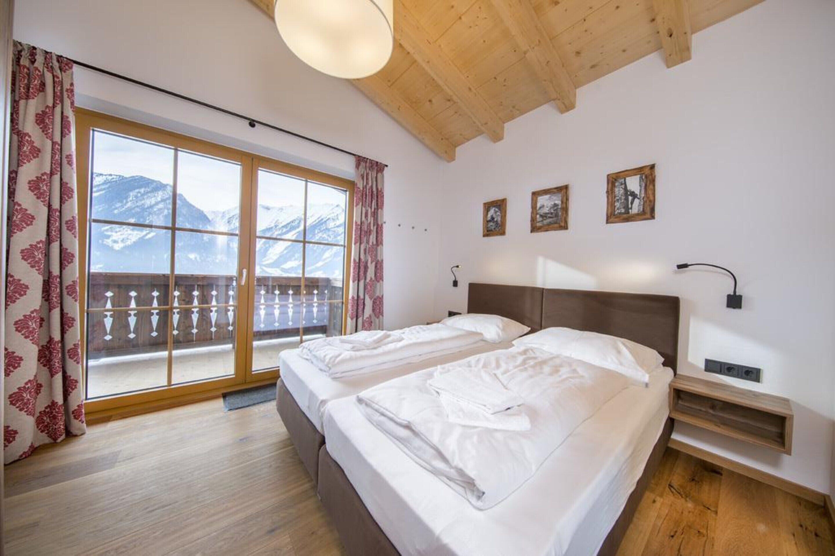 Une chambre avec deux lits, un plafond en bois, un balcon offrant une vue sur des montagnes enneigées et trois tableaux encadrés accrochés au mur.