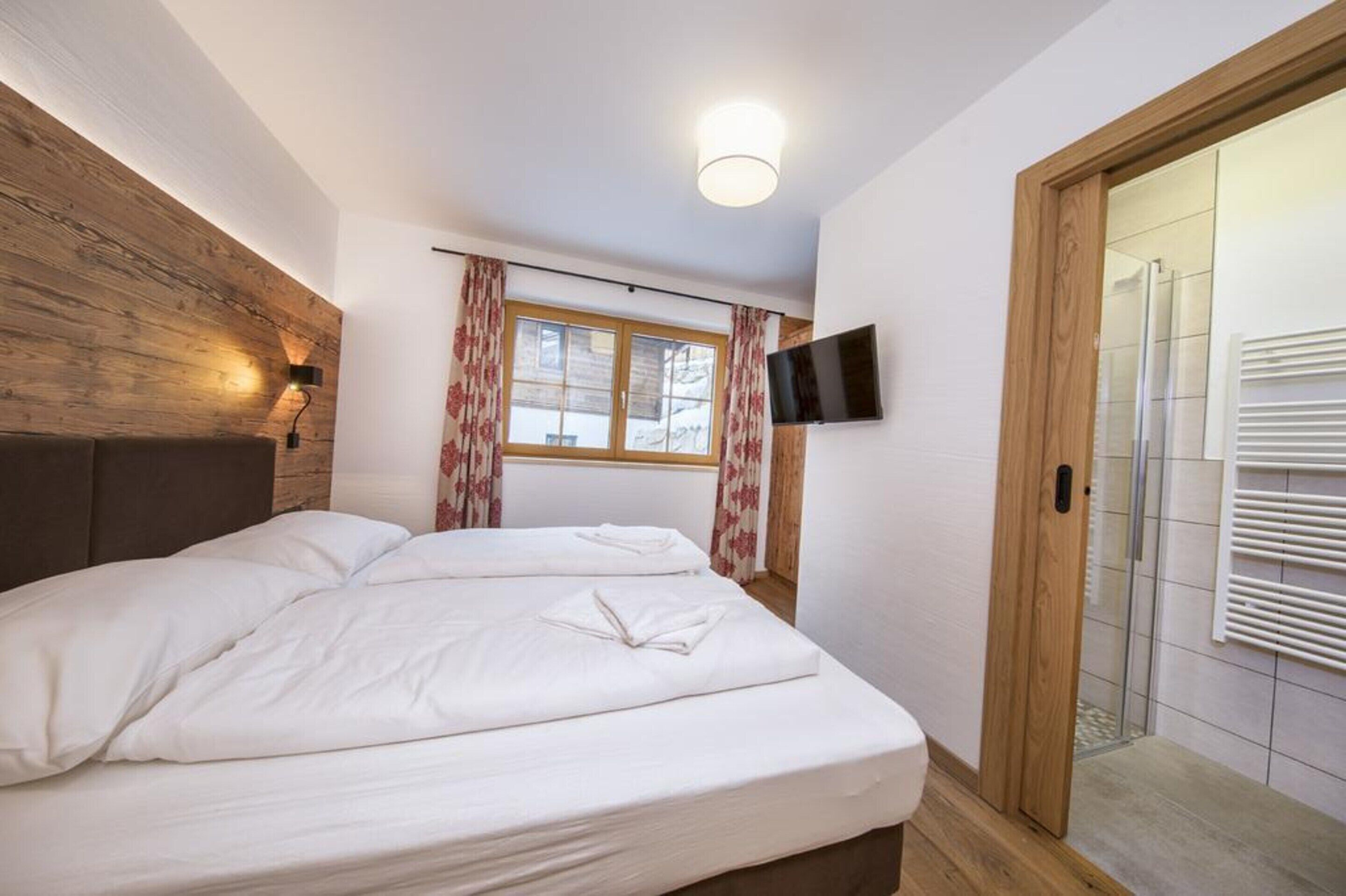 A bedroom with a wooden headboard, a bed with white linens, a window with curtains, a mounted TV, and a sliding door with a radiator.