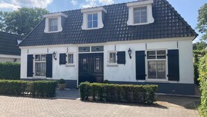 A two-story house with a dark tiled roof, white walls, and black shutters. There are two windows on the upper floor and one on the ground floor. The house has a small front garden with hedges. - Great Holiday Home at the Efteling with Heated Swimming Pool & Hot Tub (Kaatsheuvel)