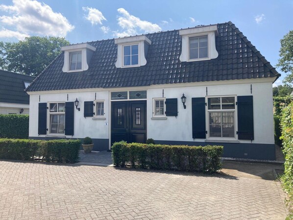 A two-story house with a dark tiled roof, white walls, and black shutters. There are two windows on the upper floor and one on the ground floor. The house has a small front garden with hedges. - Great Holiday Home at the Efteling with Heated Swimming Pool & Hot Tub (Kaatsheuvel)