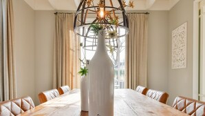 A dining table with a white vase centerpiece, surrounded by leather chairs, in a room with beige curtains and a decorative wall piece. - Great Holiday Home at the Efteling with Heated Swimming Pool & Hot Tub (Kaatsheuvel)