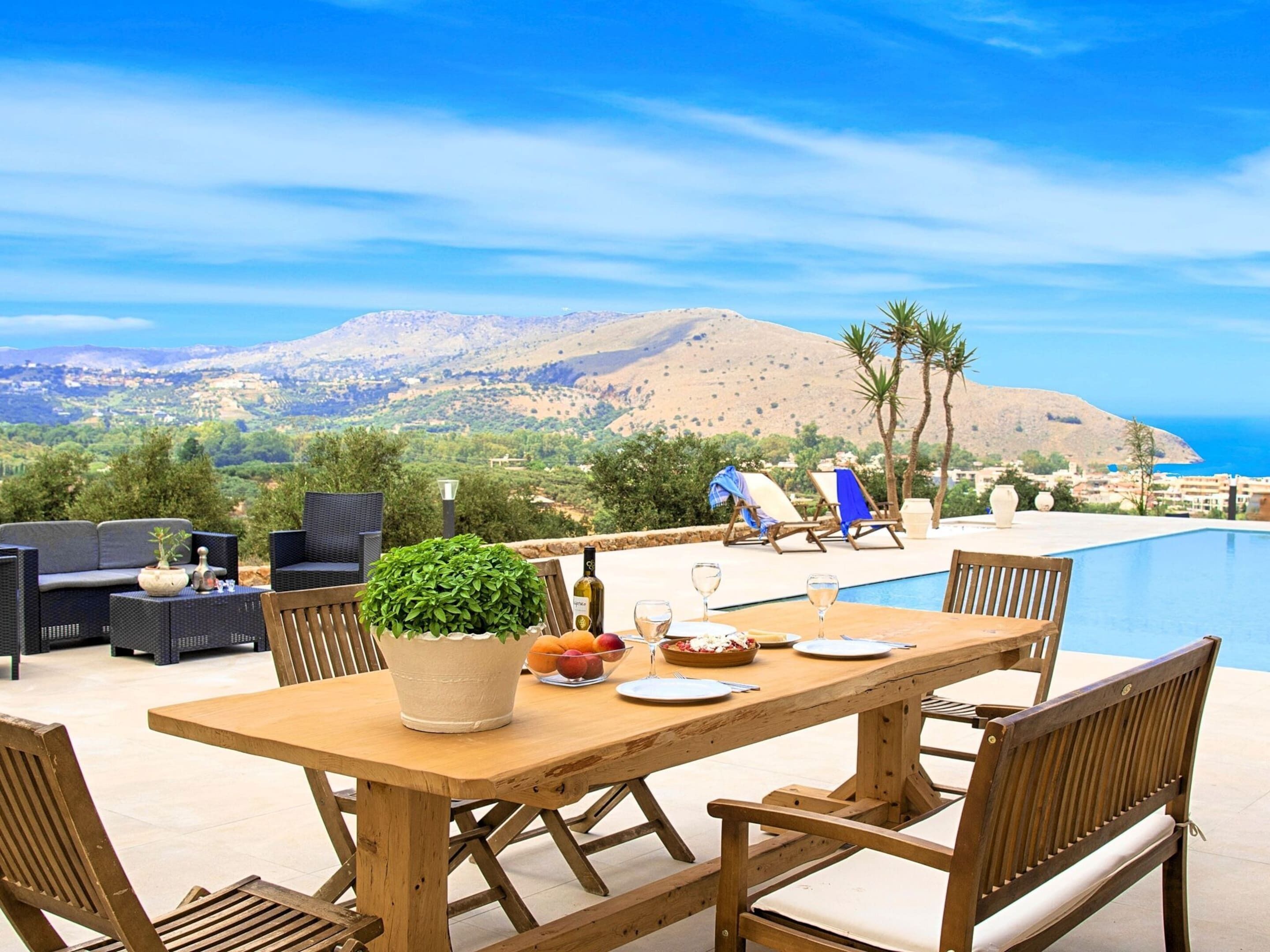 An outdoor dining area with a wooden table, chairs, and a view of a mountainous landscape and the sea.