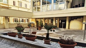 Une cour intérieure agrémentée de plantes en pot disposées en rangée, une fontaine centrale et, au fond, un bâtiment doté de grandes fenêtres et de balcons.
