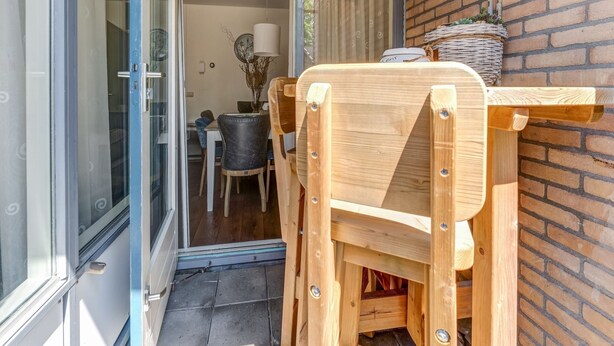 Une chaise et une table en bois se trouvent à l’extérieur d’une pièce dotée d’une porte en verre, d’un mur en briques et d’un petit espace de détente intérieur agrémenté d’une lampe et d’objets décoratifs.