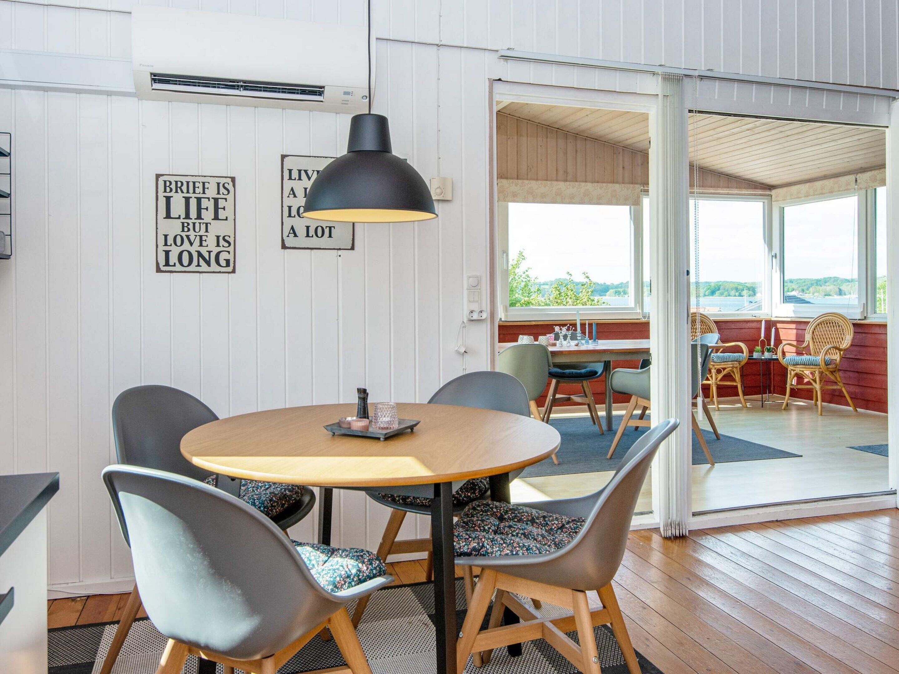Un coin repas avec une table ronde, des chaises et une suspension. Il y a deux panneaux accrochés au mur : l’un porte l’inscription « LA VIE EST COURTE, MAIS L’AMOUR EST ÉTERNEL », et l’autre, « ON VIVRA BEAUCOUP ».