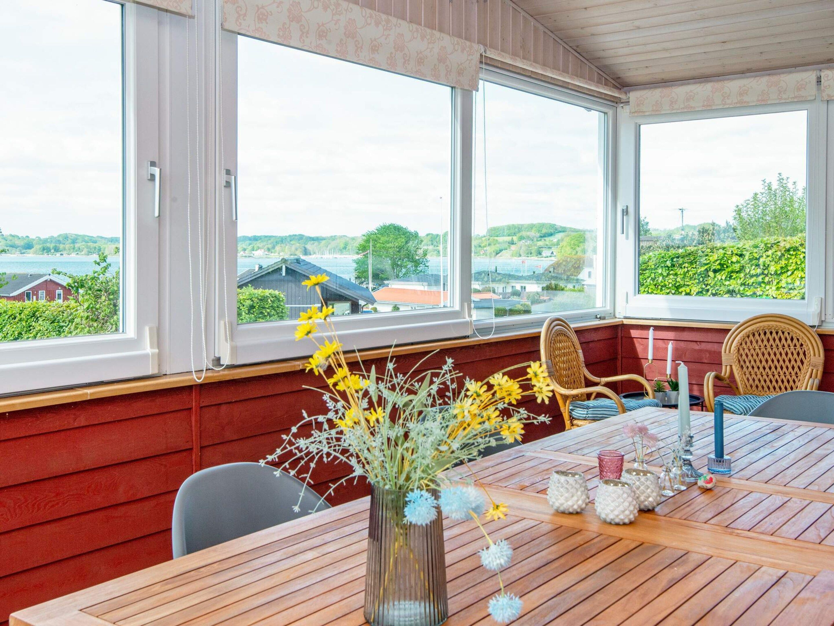 A room with a wooden table, two wicker chairs, a vase with flowers, and a view of a lake and buildings.