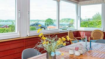 A room with a wooden table, two wicker chairs, a vase with flowers, and a view of a lake and buildings.
