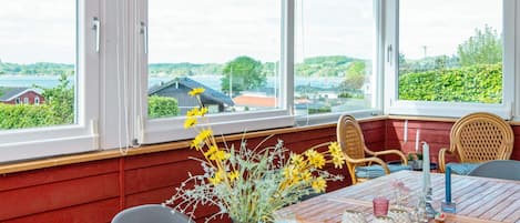 A room with a wooden table, two wicker chairs, a vase with flowers, and a view of a lake and buildings.