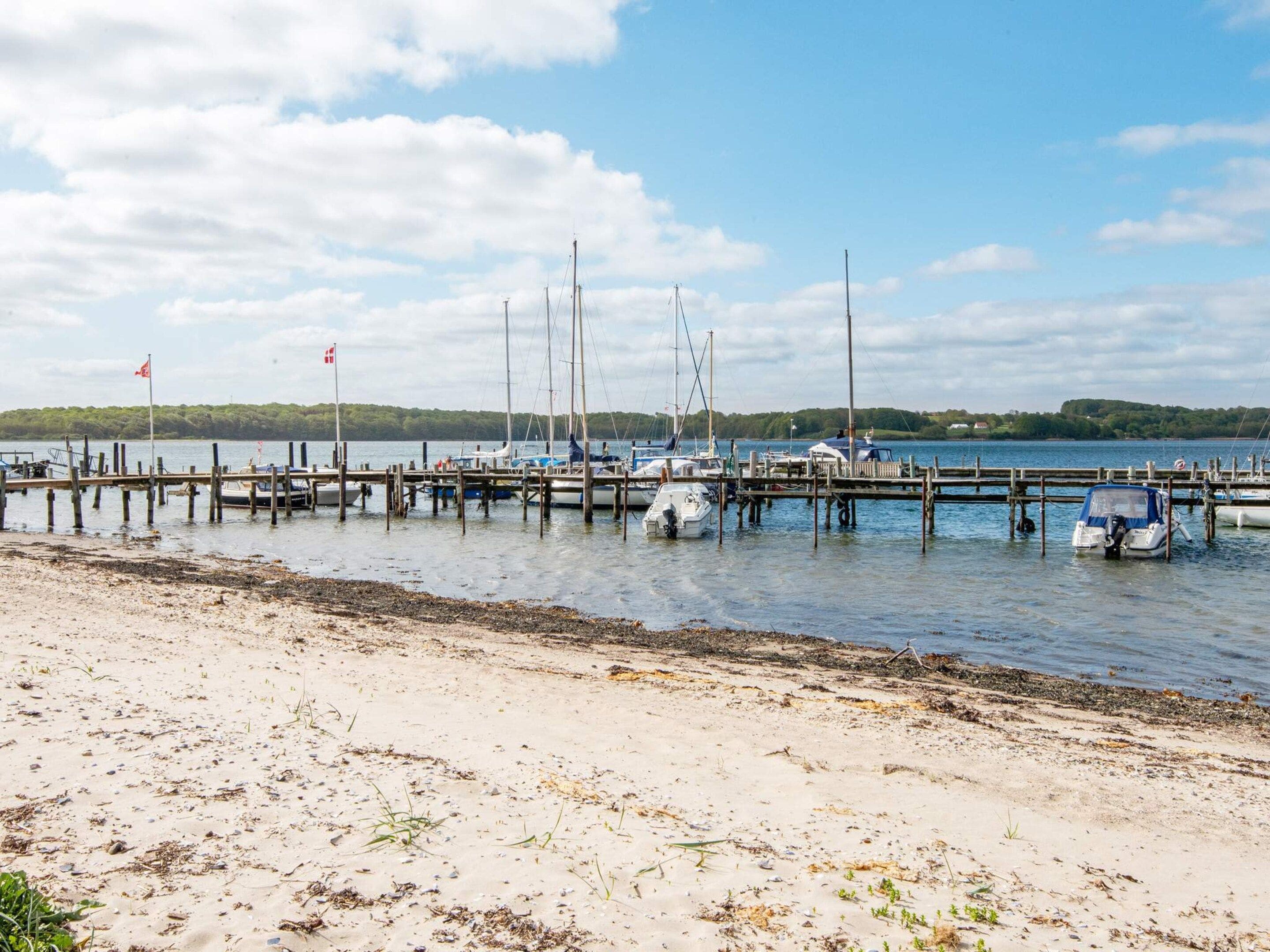 A marina with boats docked at wooden piers, a sandy beach in the foreground, and a forested shoreline in the background.