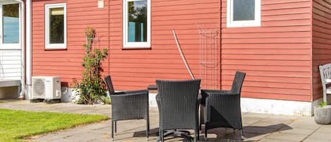 A red house with two windows, a patio with chairs, and a small garden.