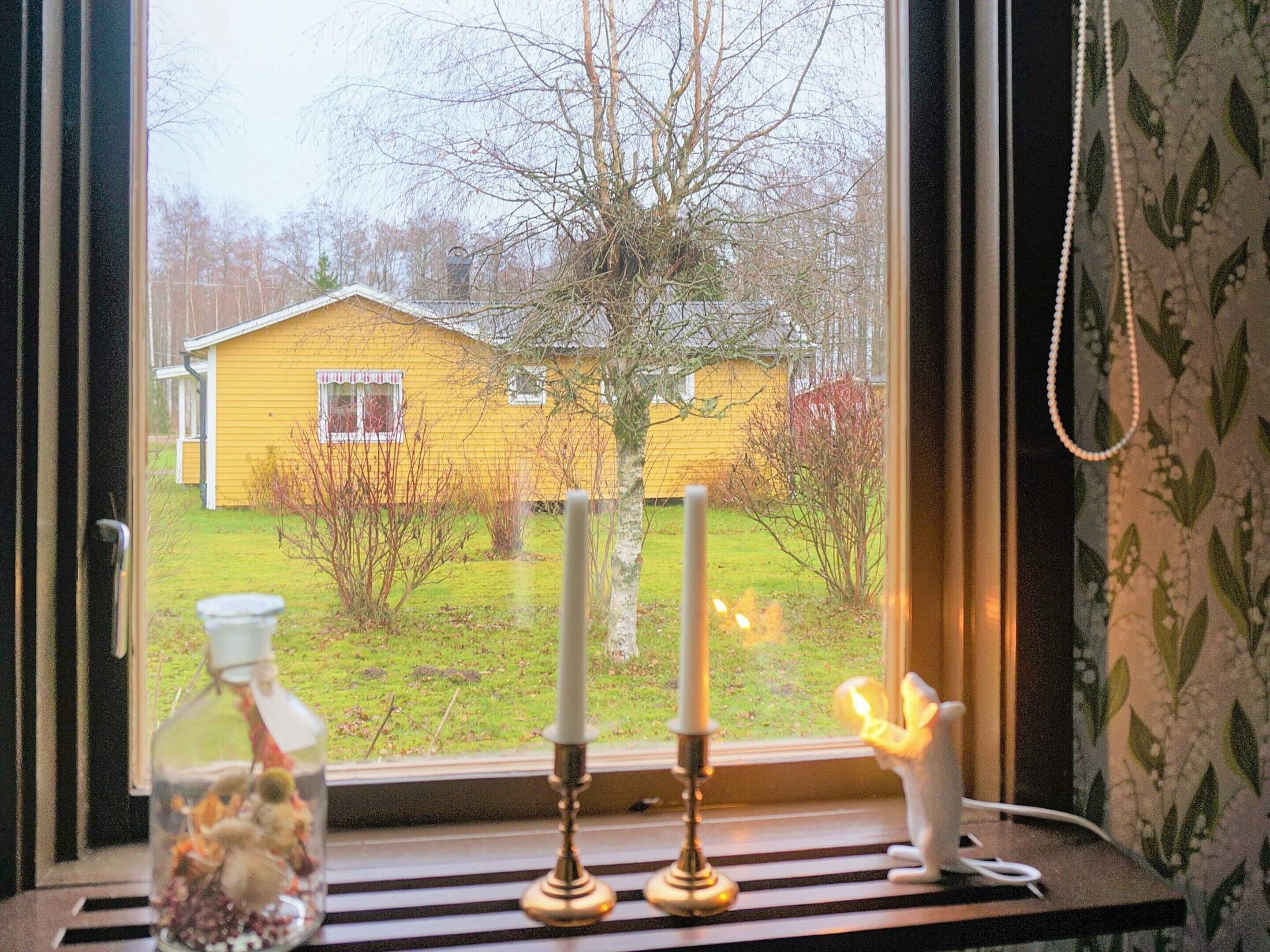 Ikkunasta avautuu näkymä keltaiseen taloon. Ikkunalautalla on kynttilänjalka ja maljakko, jossa on kynttilä.